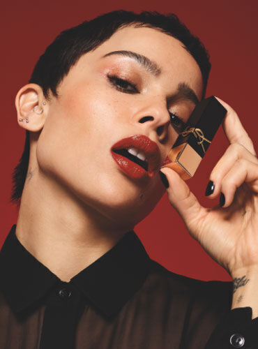 A person with short hair poses against a red background, holding a lipstick in one hand while applying it with the other. They wear bold red lipstick and a sheer black shirt.