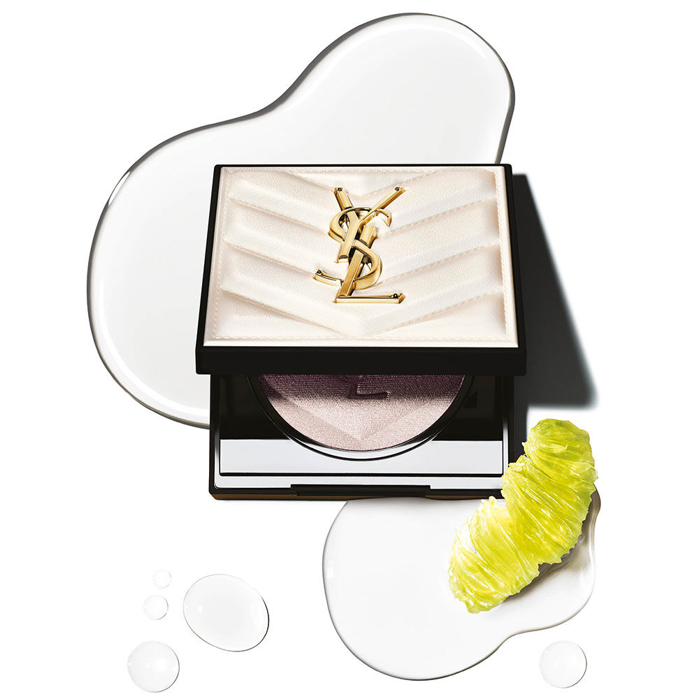 A compact of YSL highlighter with a shiny gold logo, resting on a white background with decorative glossy drops and a slice of lime.