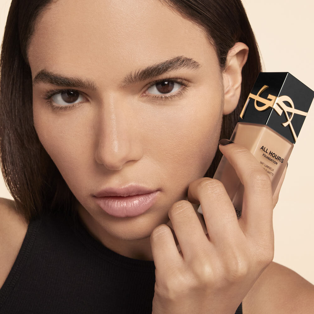 A close-up of a model holding a bottle of foundation, showing a natural makeup look with smooth skin and neutral tones. The model has dark, straight hair and a serious expression.