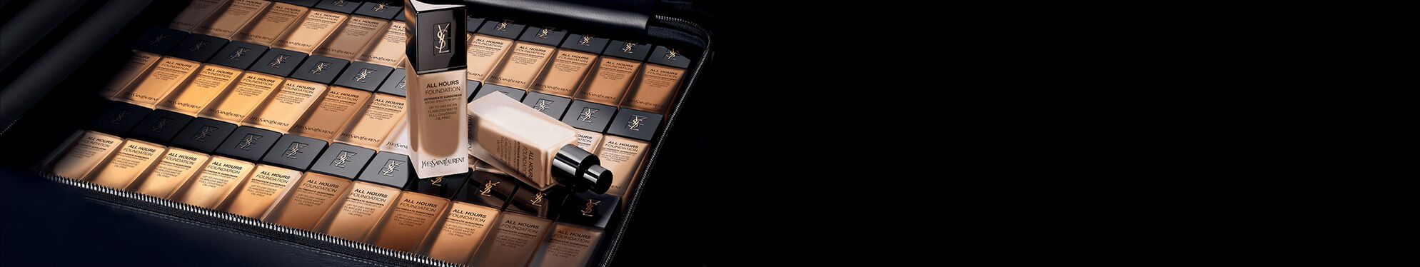 A collection of foundation bottles arranged neatly inside a black case, showcasing various shades of the product. The scene has a sleek, modern aesthetic with a dark background.