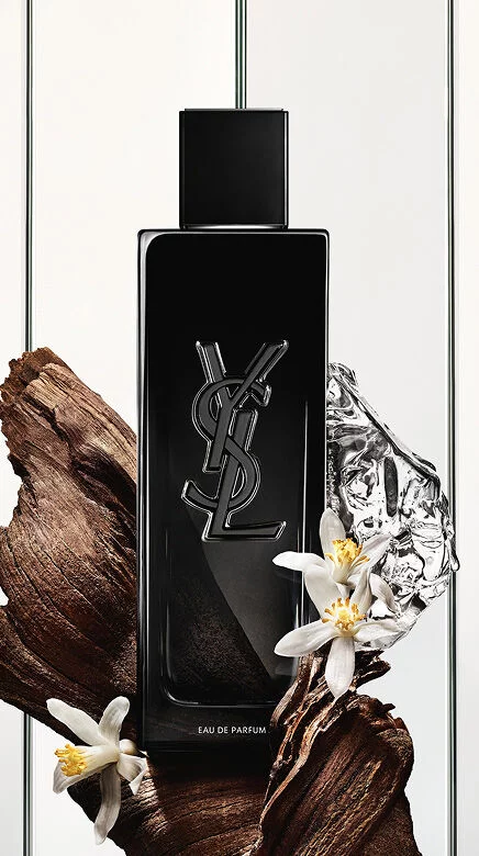 Eine schwarze Yves Saint Laurent Eau de Parfum Flasche mit dem YSL-Logo, umgeben von dunklen Holzstucken, zarten weißen Bluten und einem zerknitterten silbernen, reflektierenden Objekt.