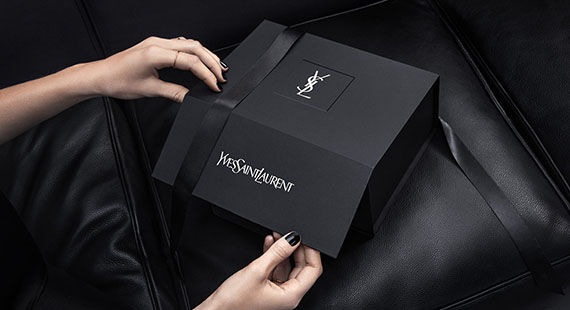 A pair of hands is opening a sleek black box with a ribbon, featuring the Yves Saint Laurent logo on top, placed on a dark leather surface.