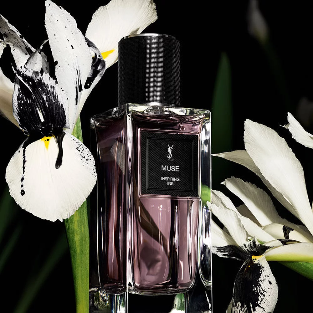 Une bouteille de parfum rectangulaire etiquetee "MUSE INSPIRING INK" de YSL, entouree d'elegantes iris blanches et noires.