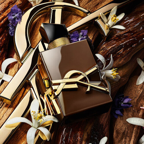 YSL Libre Parfumflakon, umgeben von goldenen Elementen, weißen Orangenbluten und lila Blumen auf Holzgrund.