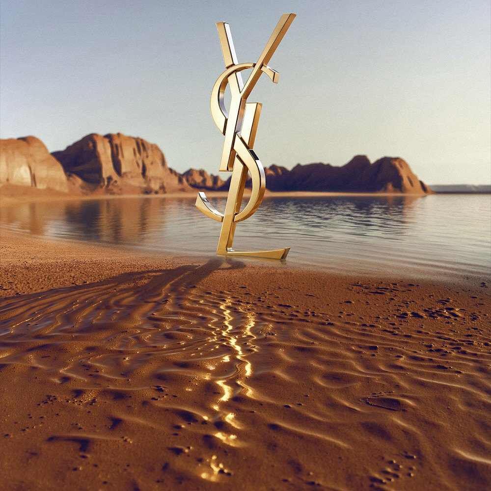 Ein großes, ikonisches 3D-YSL-Logo (Yves Saint Laurent) steht an einem Wustenstrand. Das polierte goldene Monogramm spiegelt das Sonnenlicht wider und ist vor einer ruhigen Wasserflache mit dramatischen roten Felsformationen und Wustenbergen im Hintergrund unter einem klaren Himmel positioniert.