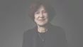 Un portrait rapproche d'une femme souriante aux cheveux bruns courts, vetue d'un costume sombre et d'un collier delicat, sur un fond sombre.