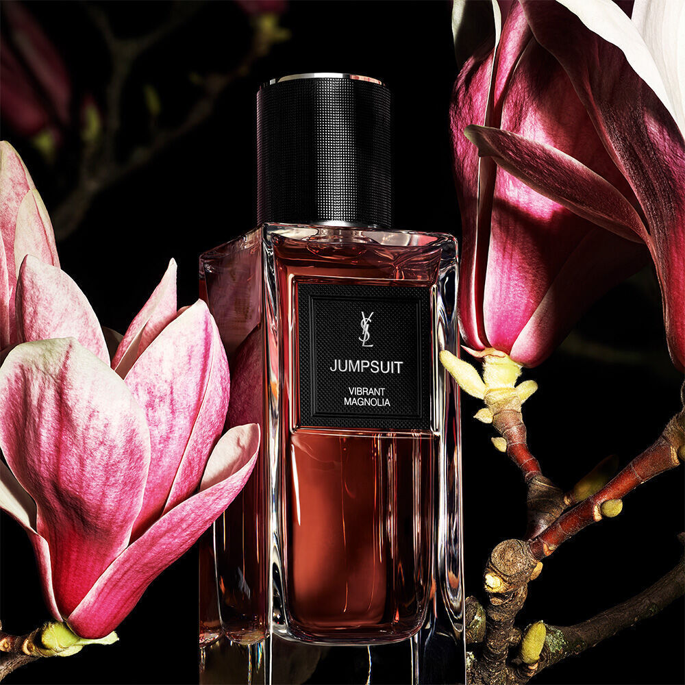 Une bouteille de parfum brun-rougeatre etiquetee "JUMPSUIT VIBRANT MAGNOLIA" de YSL, placee sur un fond sombre avec de grands petales de magnolia roses.