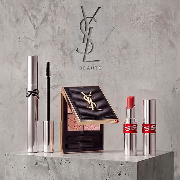 Werbeprasentation von YSL Beaute Make-up-Produkten, darunter Mascara, eine gesteppte Lidschattenpalette und Lippenstifte vor einem Betonhintergrund.
