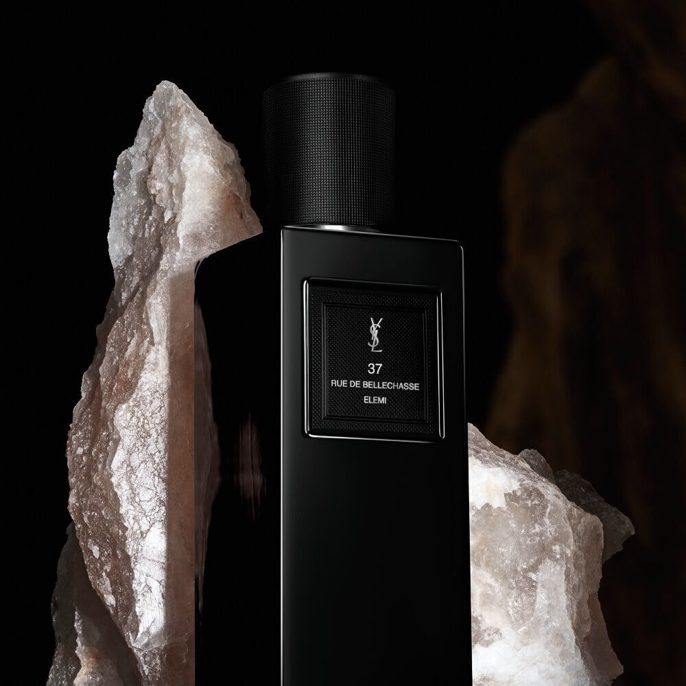 Une elegante bouteille de parfum noire et rectangulaire etiquetee "37 RUE DE BELLECHASSE ELEMI" de YSL, encadree par de grandes formations de cristaux blancs et bruts.