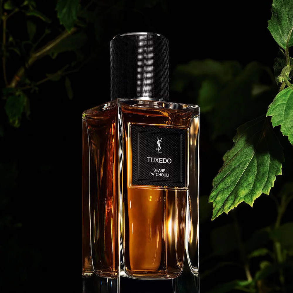 Une bouteille de parfum aux tons dores etiquetee "TUXEDO SHARP PATCHOULI" de YSL, avec un doux feuillage vert en arriere-plan naturel.