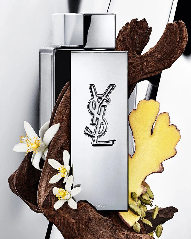 YSL Y L'ABSOLU Parfumflakon umgeben von dunklem Holz, weißen Orangenbluten, Ingwer und Kardamomkapseln.