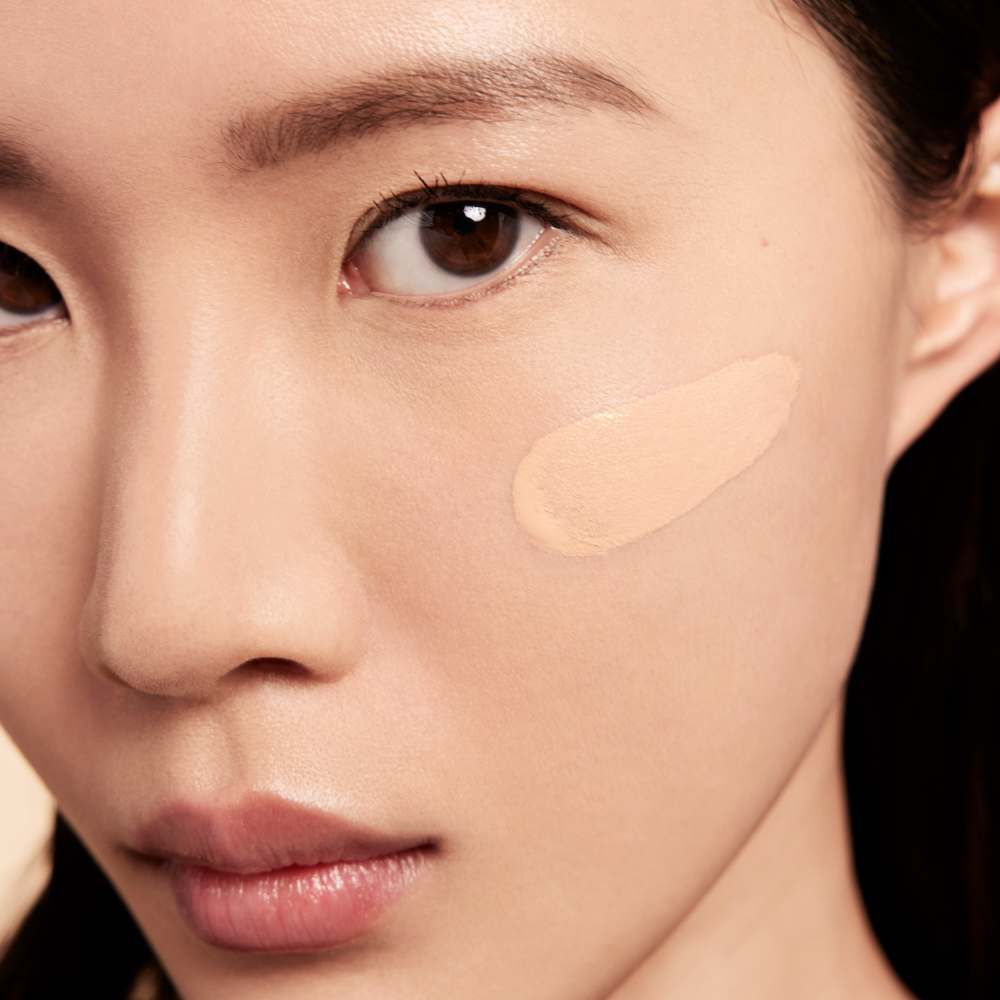 A close-up of a person's face with a light peach-colored foundation applied to one cheek, showcasing natural skin texture and features.