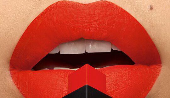 Close-up of a pair of lips painted in bright red lipstick, partially open, with a geometric red and black lipstick tube positioned below.