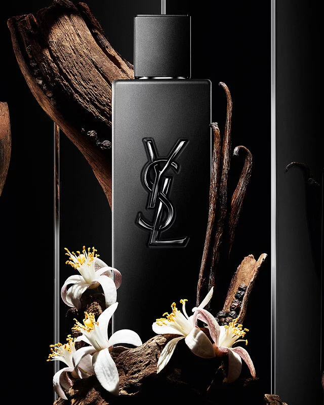 Schwarzer YSL-Parfumflakon, umgeben von Holz, Vanilleschoten und weißen Bluten.