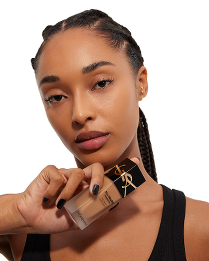 A woman with braided hair holds a bottle of YSL foundation close to her face, showcasing her flawless complexion and natural makeup.