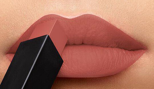 Close-up of a pair of lips with a smooth matte finish, as a lipstick is being applied. The lipstick shade is a soft nude tone, and the packaging is sleek and rectangular.