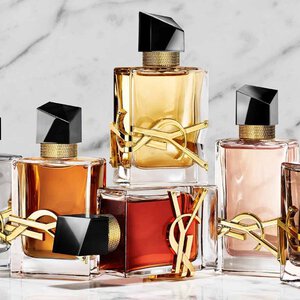 Une main tient un flacon de parfum YSL rouge a cote de deux autres fragrances sur un socle en pierre.