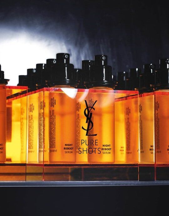 Rangee de flacons de serum Yves Saint Laurent Pure Shots Night Reboot de couleur ambre avec des pompes noires. Le logo YSL est proeminent sur chaque flacon, qui repose sur une surface reflechissante devant un fond sombre et fume, mettant en valeur la ligne de produits.