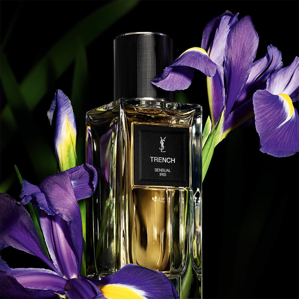 Une bouteille de parfum aux tons dores etiquetee "TRENCH SENSUAL IRIS" de YSL, mise en evidence a cote de vibrantes iris violettes.