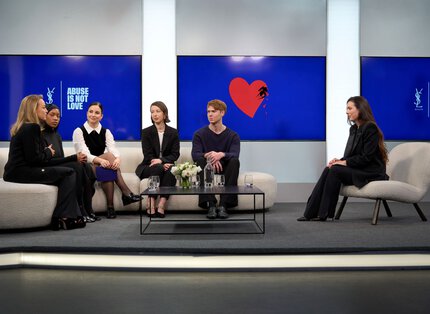Podiumsdiskussion mit sechs Sprechern (vier Frauen, zwei Manner) auf Sofas sitzend, vor großen blauen Bildschirmen mit Botschaften, in einem modernen Raum.