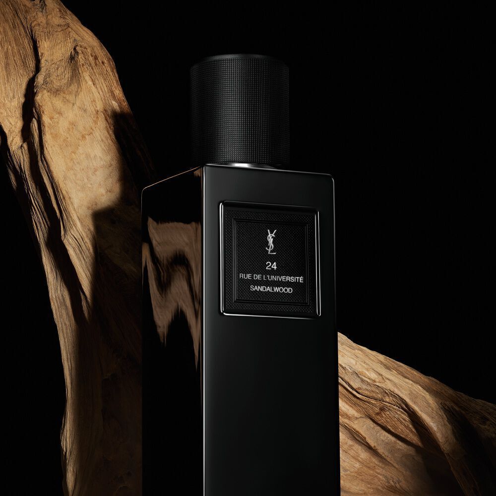 Une elegante bouteille de parfum noire et rectangulaire etiquetee "24 RUE DE L'UNIVERSITE SANDALWOOD" de YSL, positionnee sur un fond de bois naturel et rustique.