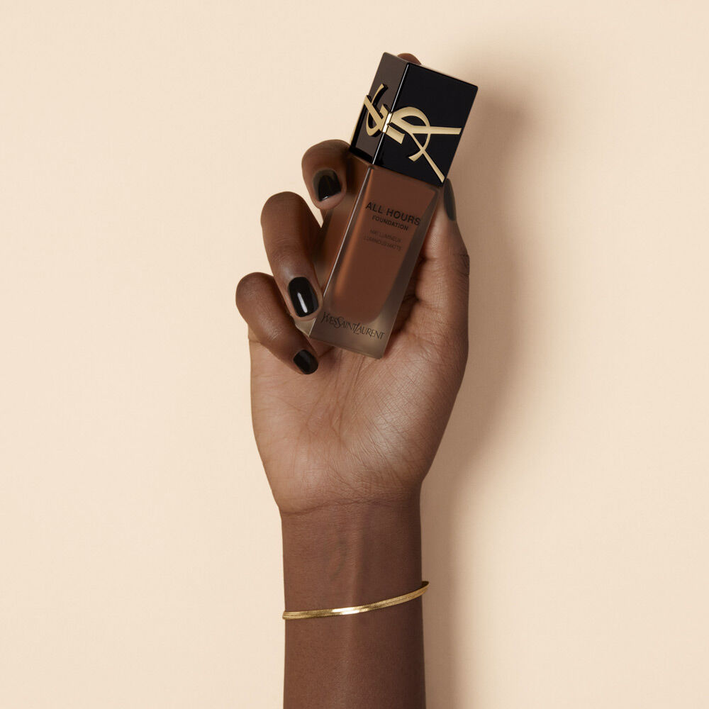 A hand with dark skin is holding a bottle of YSL All Hours Foundation in a warm brown shade, with a sleek, elegant black and gold design. The background is a soft beige color.