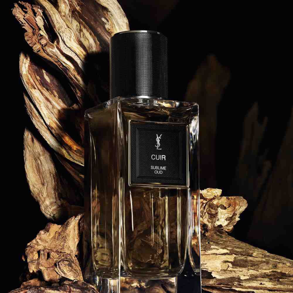 Une bouteille de parfum teintee de brun etiquetee "CUIR SUBLIME OUD" de YSL, entouree de morceaux de bois vieilli et texture.
