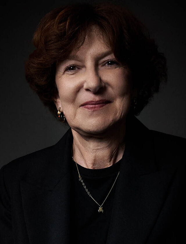 Une femme souriante aux cheveux bruns courts, vetue d'un costume sombre et d'un collier delicat, est assise sur une chaise en cuir fonce.