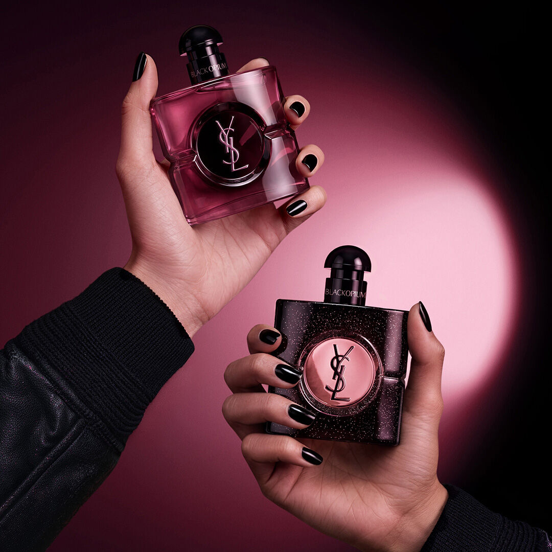 Deux mains tenant des flacons de parfum Yves Saint Laurent Black Opium sur un fond rose fonce et noir.