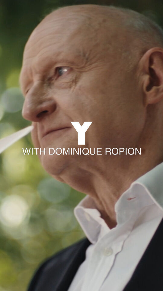 Un homme chauve, vetu d'un costume noir et d'une chemise blanche, porte a son nez plusieurs bandes de papier blanc, couramment utilisees pour tester les parfums, comme s'il evaluait une odeur. Il a l'air contemplatif, sur un fond vert et flou de feuillage et d'arbres. L'image est recouverte d'un texte blanc de grande taille "Y" et d'un texte plus petit en dessous, "AVEC DOMINIQUE ROPION".
