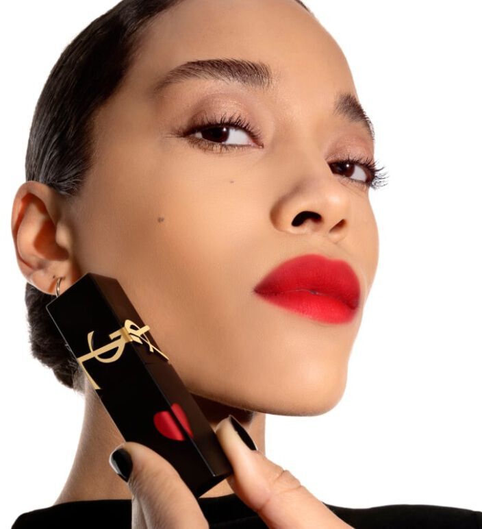 A person exemplifies confidence while holding a black lipstick tube near their face, displaying bold red lips and striking eye makeup against a white background.