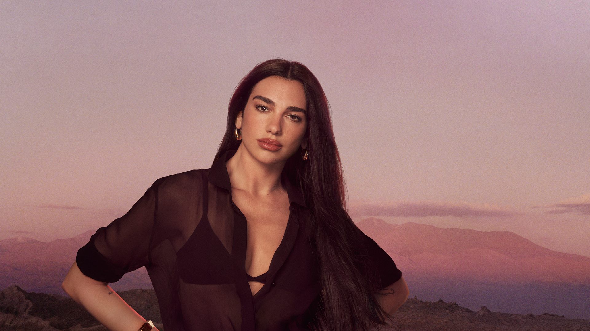 Dua Lipa posiert in einem schwarzen, transparenten Hemd vor einer bergigen Gebirgslandschaft bei Sonnenuntergang.