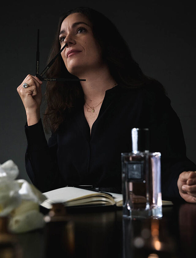 Une femme en chemise noire tient une touche a parfum, levant les yeux pensivement, avec un carnet, une bouteille de parfum et des fleurs sur la table.