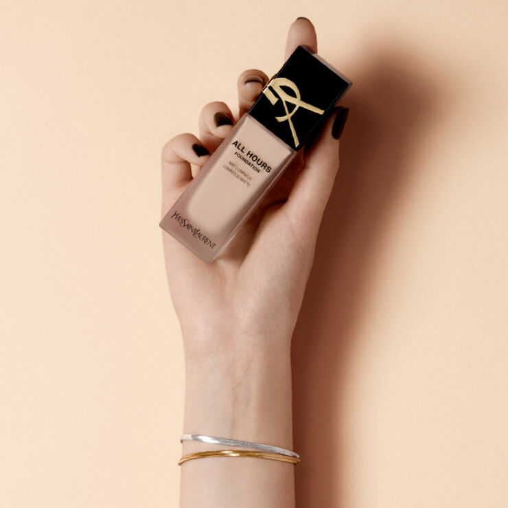 A hand holding a bottle of Yves Saint Laurent All Hours foundation against a light peach background.