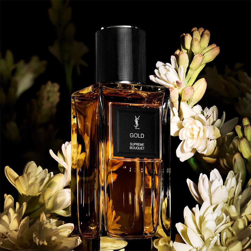 Une bouteille de parfum aux tons dores etiquetee "GOLD SUPREME BOUQUET" de YSL, richement entouree d'un bouquet de fleurs de tubereuse blanches.
