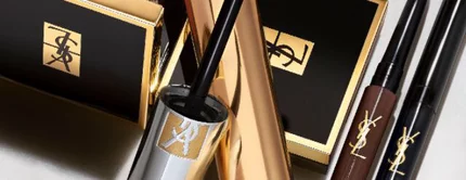 A collection of beauty products arranged on a marble surface, including mascara, a brown eyeshadow compact, and eyeliners, all featuring elegant packaging.