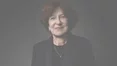 Un portrait rapproche d'une femme souriante aux cheveux bruns courts, vetue d'un costume sombre et d'un collier delicat, sur un fond sombre.
