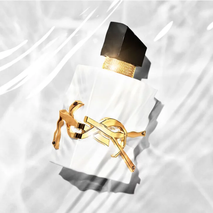 Ein eleganter Parfumflakon mit schwarzem Deckel und goldener Verzierung, der auf einem hellen, reflektierenden Untergrund liegt.