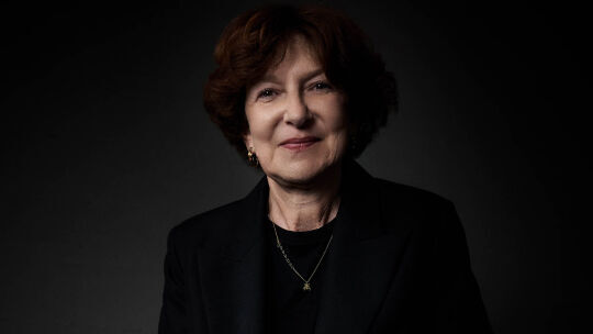 Un portrait rapproche d'une femme souriante aux cheveux bruns courts, vetue d'un costume sombre et d'un collier delicat, sur un fond sombre.