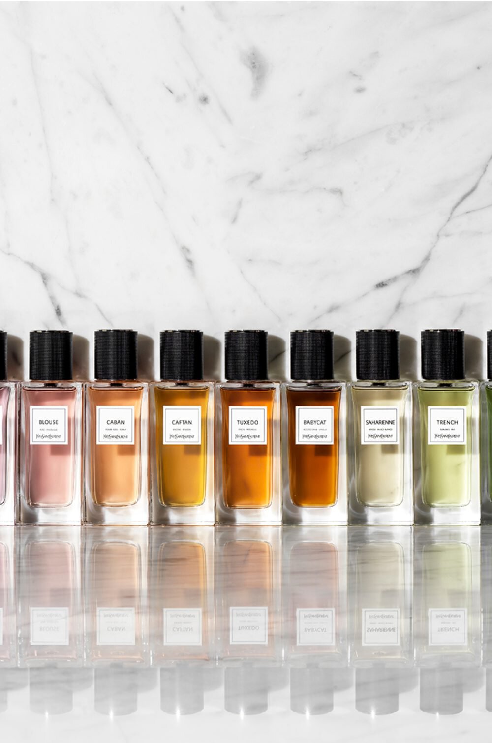 A row of 12 elegantly designed perfume bottles on a marble surface, each with a labeled black cap and varying shades of liquid, from deep red to green.