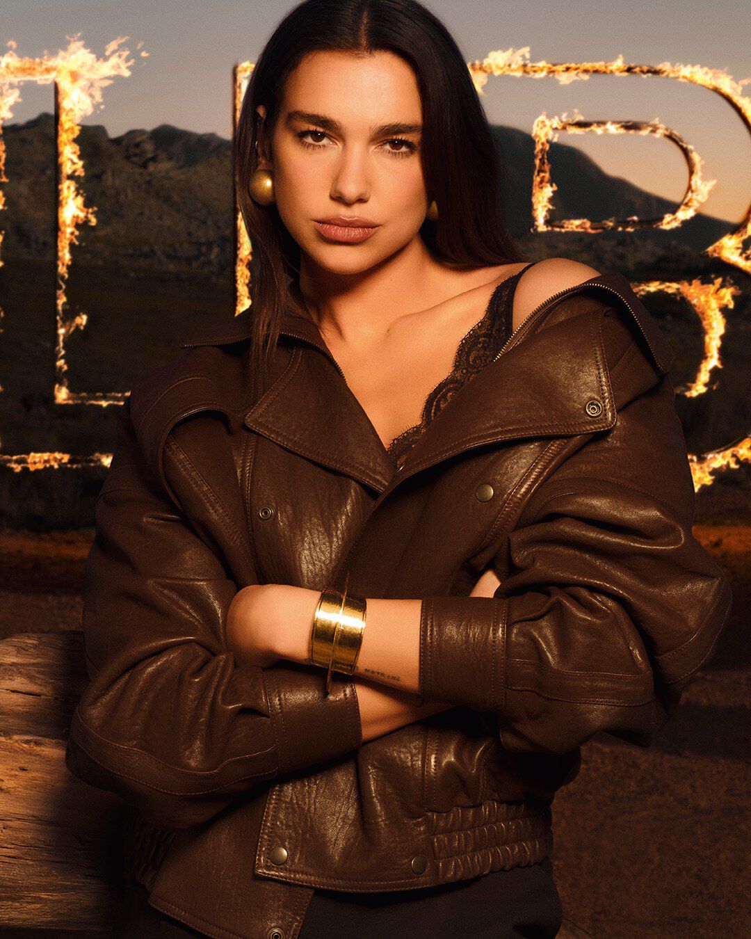 Dua Lipa in einer braunen Lederjacke vor großen, flammenden LIBRE Buchstaben in einer rauen Wustenlandschaft.