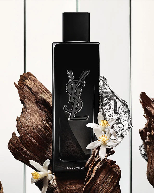 Eine schwarze Yves Saint Laurent Eau de Parfum Flasche mit dem YSL-Logo, umgeben von dunklen Holzstucken, zarten weißen Bluten und einem zerknitterten silbernen, reflektierenden Objekt.