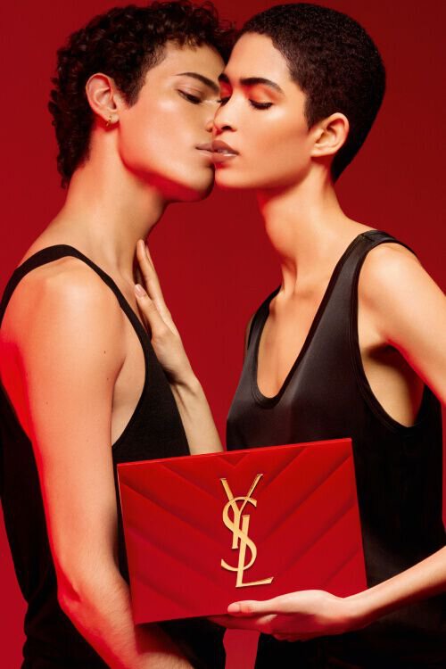 Deux modeles aux cheveux courts s'embrassent doucement, tenant un sac rouge avec le logo YSL. L'arriere-plan est rouge uni, creant une ambiance chaleureuse et intime.