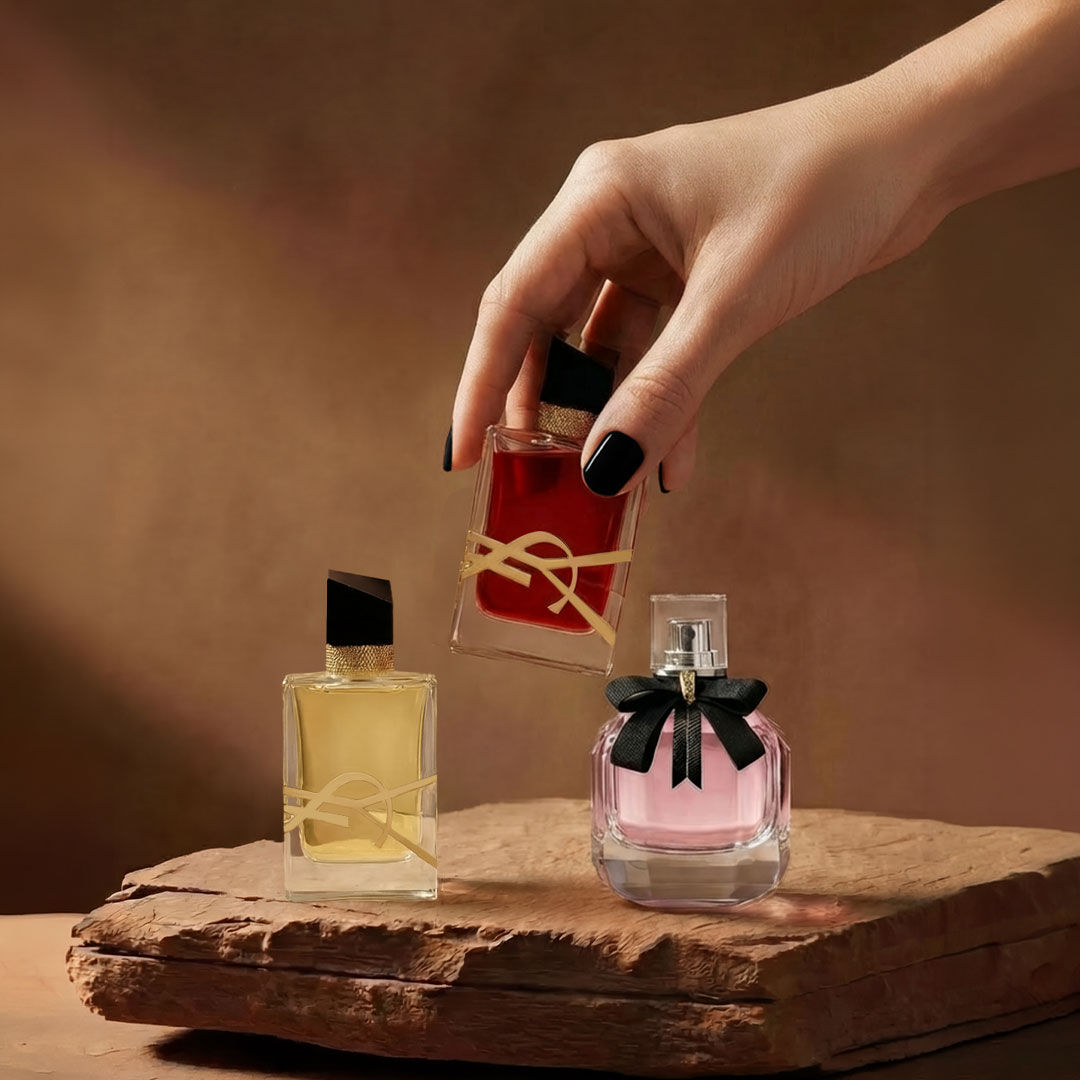 Une main tient un flacon de parfum YSL rouge a cote de deux autres fragrances sur un socle en pierre.