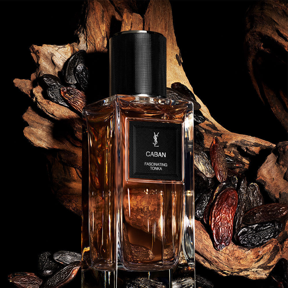 Une bouteille de parfum aux tons ambres etiquetee "CABAN FASCINATING TONKA" de YSL, entouree de feves tonka et d'un morceau de bois flotte noueux.