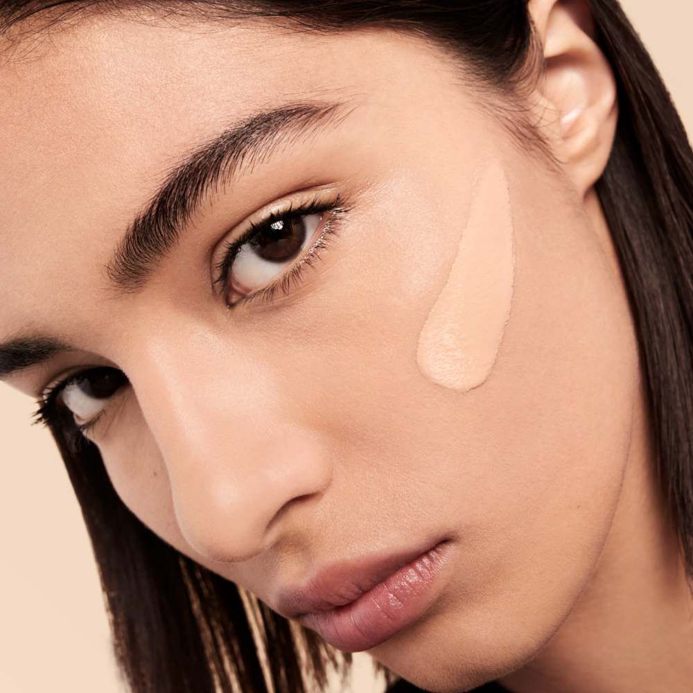 A close-up of a person's face, featuring a smooth application of foundation on one cheek. The individual's expression is neutral, and they have dark hair and eyebrows.