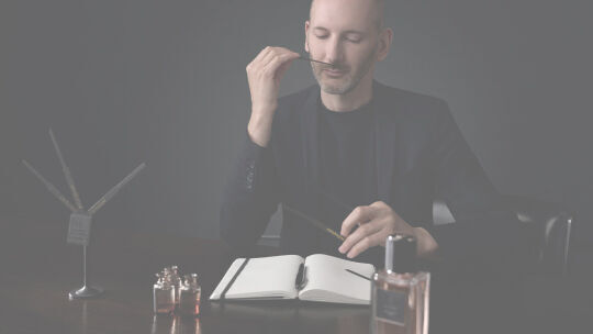 Un parfumeur en costume sombre sent attentivement une touche a parfum dans un cadre faiblement eclaire.