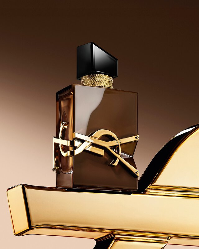 Flacon Libre Santal Couture brun glace refletant la lumiere, pose sur une grande Cassandre YSL doree.