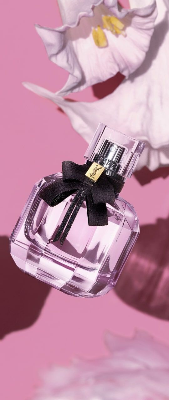 A clear glass perfume bottle with a black bow rests against a pink background, accompanied by soft white flowers.