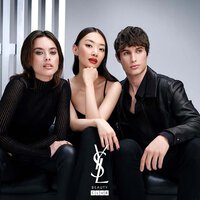 A group of three stylish models poses together in a minimalist setting. They convey a sense of confidence and fashion, with the logo "YSL BEAUTY CLUB" featured prominently at the bottom.